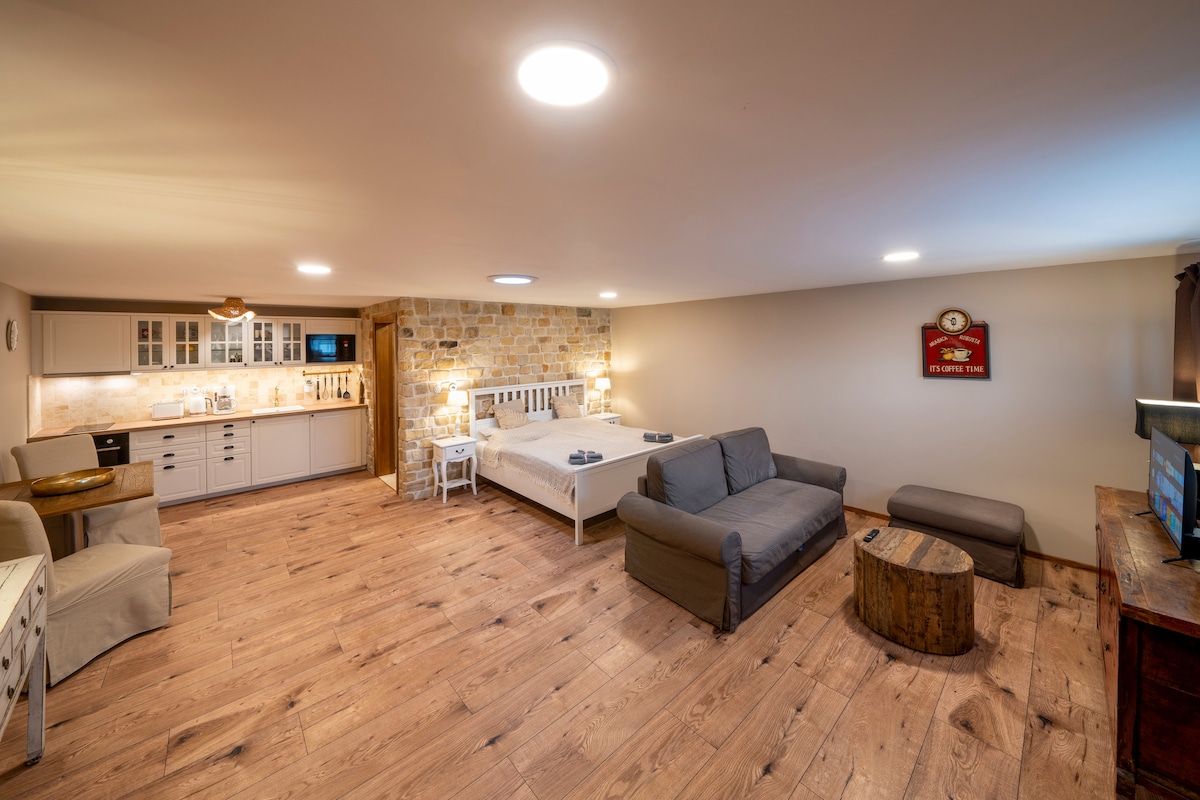 The spacious studio includes a fully equipped kitchenette with light-colored cabinetry, a cozy seating area with a gray sofa, and a large double bed positioned alongside rustic stone walls. Warm wood floors contribute to the inviting atmosphere throughout the room.