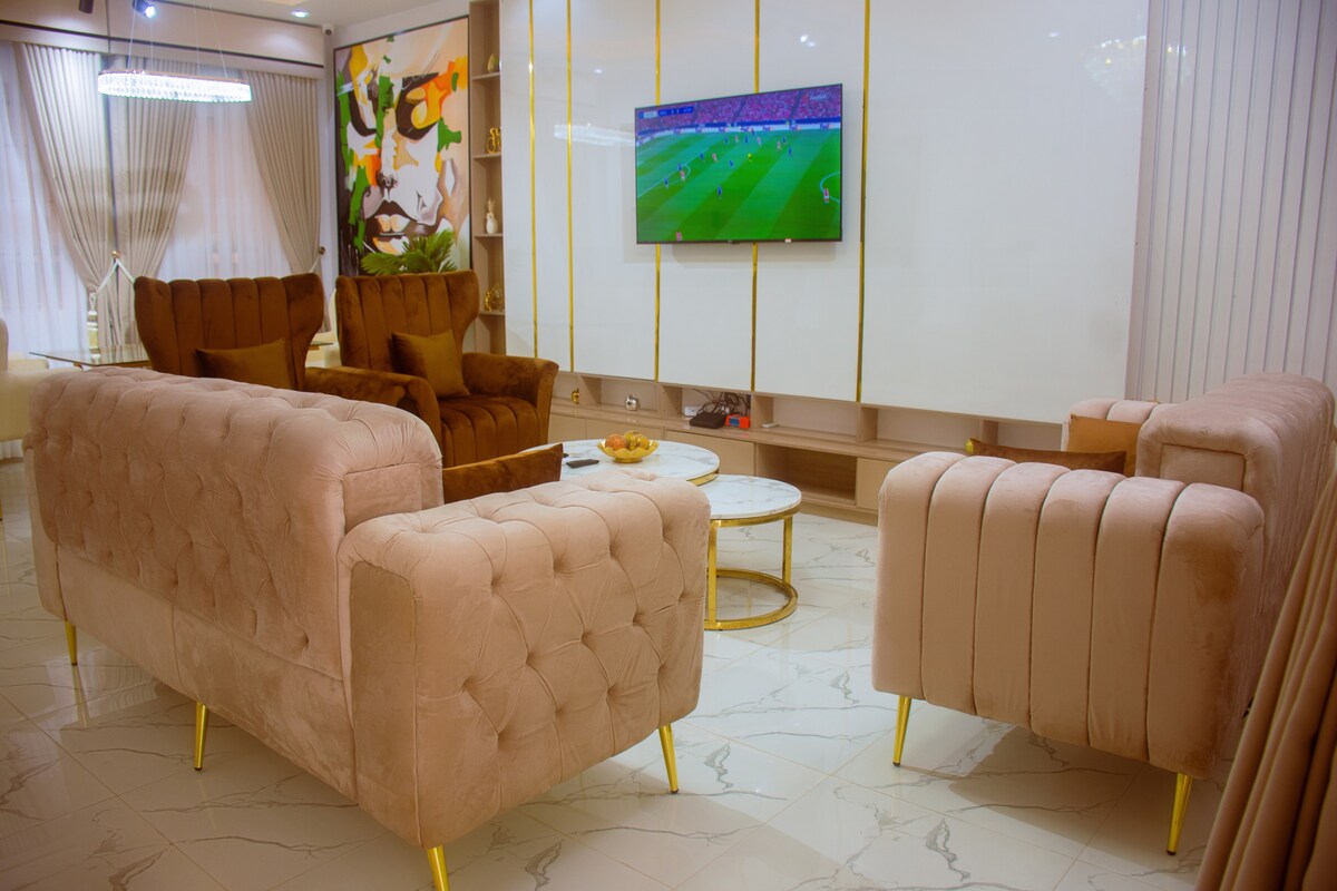 A living space features two plush sofas with a tufted design, positioned around a circular marble coffee table. A flat-screen television is mounted on a wall, displaying an image. Soft lighting enhances the modern decor, while large windows allow natural light to fill the room.