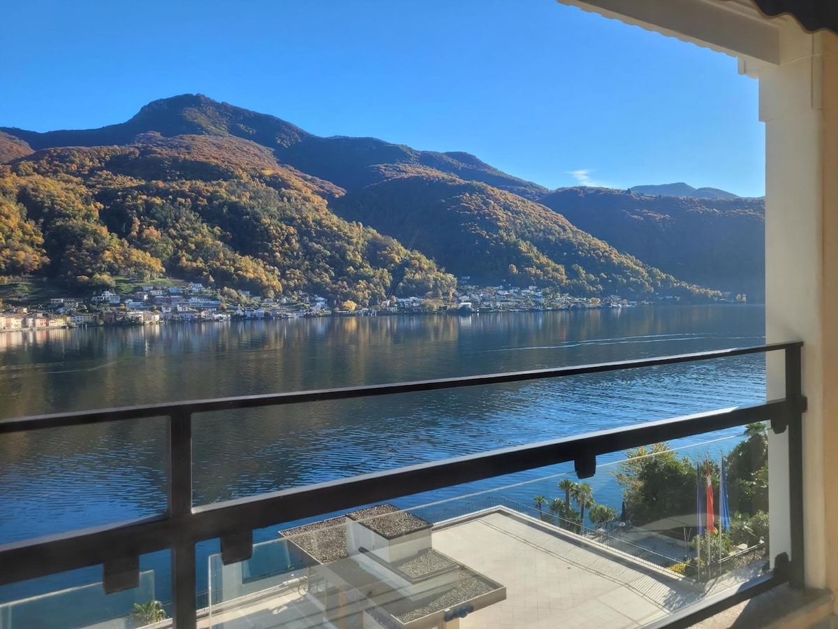 A panoramic view of Lake Lugano is captured from the balcony, framed by mountains adorned with autumn foliage. The clear blue sky reflects on the water's surface, enhancing the serene ambiance of the surrounding landscape.