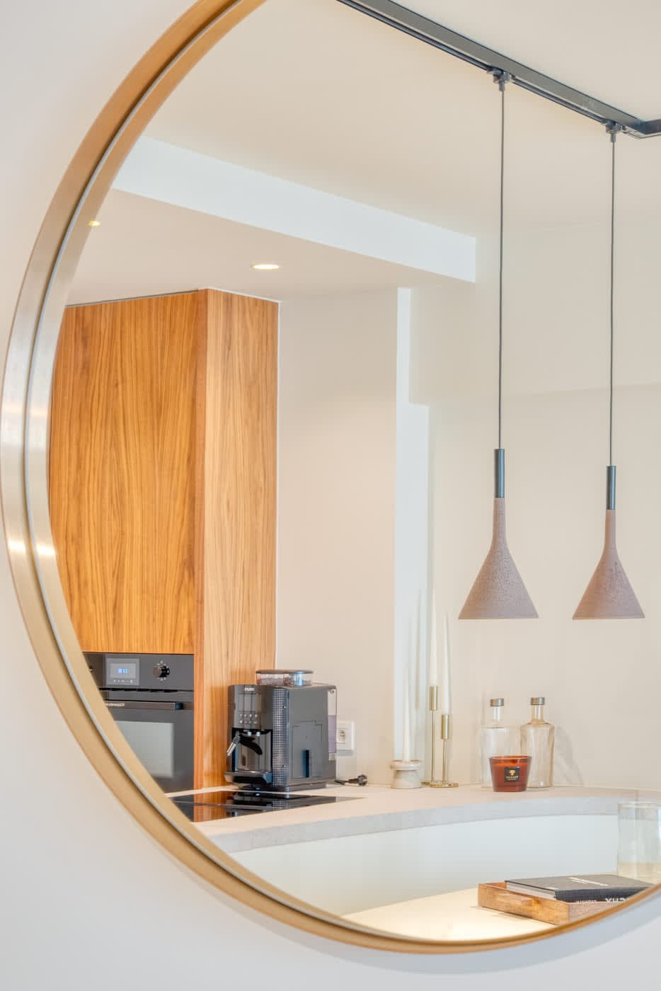 A modern kitchen space is reflected in a circular mirror, showcasing warm wood cabinetry and sleek appliances. Two pendant lights hang gracefully above the countertop, complementing the minimalist design, while glass containers are positioned neatly in the background.