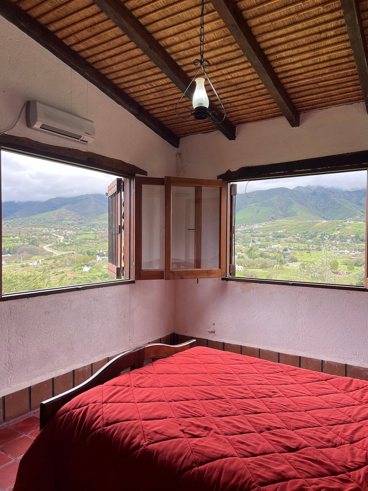 A cozy bedroom features a red quilted bedspread on a spacious bed. Two large windows offer views of the surrounding mountains and valleys, allowing natural light to fill the room. The ceiling is adorned with wooden beams, complemented by a hanging light fixture.