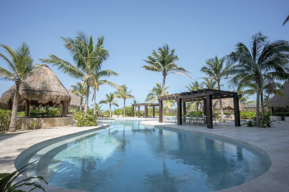 An outdoor pool is surrounded by palm trees and tropical landscaping. Thatched-roof cabanas offer shaded seating areas beside the water. Clear blue skies create a bright atmosphere, complementing the inviting pool area, which features a smooth stone deck and well-maintained greenery.