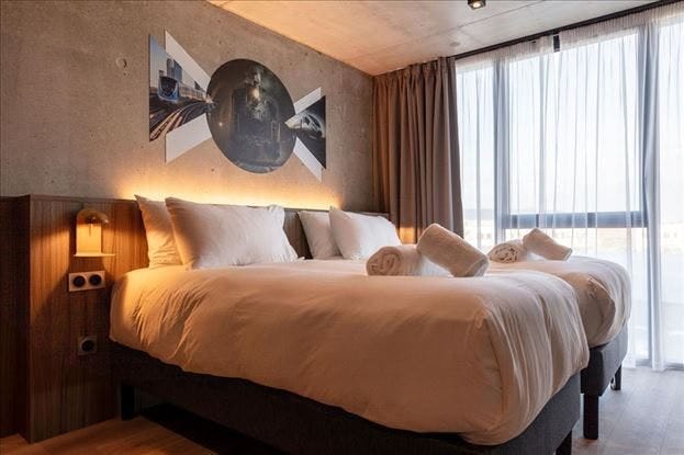 A light-filled room features two spacious beds, each adorned with soft white linens and neatly rolled towels. A contemporary mural is displayed above the beds, while sheer curtains allow natural light to filter in, enhancing the room's inviting ambiance.