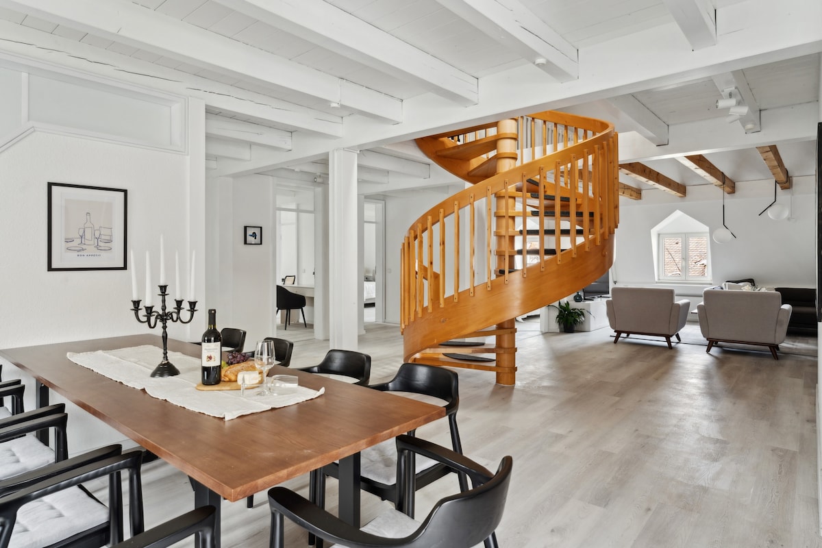 An elegant dining area features a wooden table surrounded by black chairs, set with candles and a bottle of wine. A spiral staircase made of wood leads to the upper levels, while bright natural light fills the open space, highlighting the light-colored flooring and exposed wooden beams.