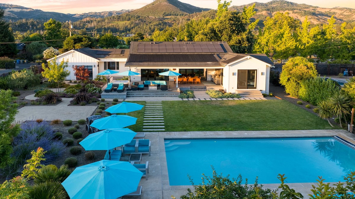 An aerial view showcases the expansive yard featuring a rectangular saltwater pool, complemented by lounge chairs and umbrellas in turquoise. The modern home is positioned to highlight large glass doors opening to the outdoor area, surrounded by lush gardens and scenic views of rolling hills.