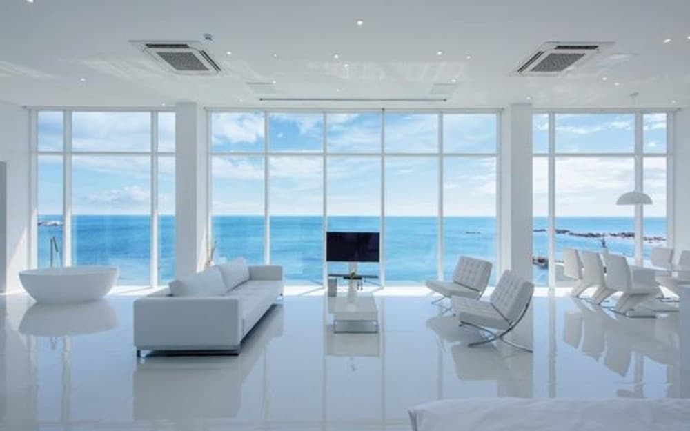 A bright and airy living area features expansive floor-to-ceiling windows that offer unobstructed ocean views. A sleek white sofa and contemporary chairs are positioned around a glass coffee table, while a TV stands against a wall. Soft light highlights the glossy surfaces.