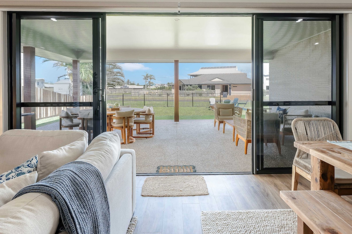 The image reveals an open indoor living space with large sliding glass doors leading to an outdoor patio area. The patio features a dining set and lounge seating, surrounded by grass and palm trees. Natural light brightens the space, creating a seamless transition between indoor and outdoor living.
