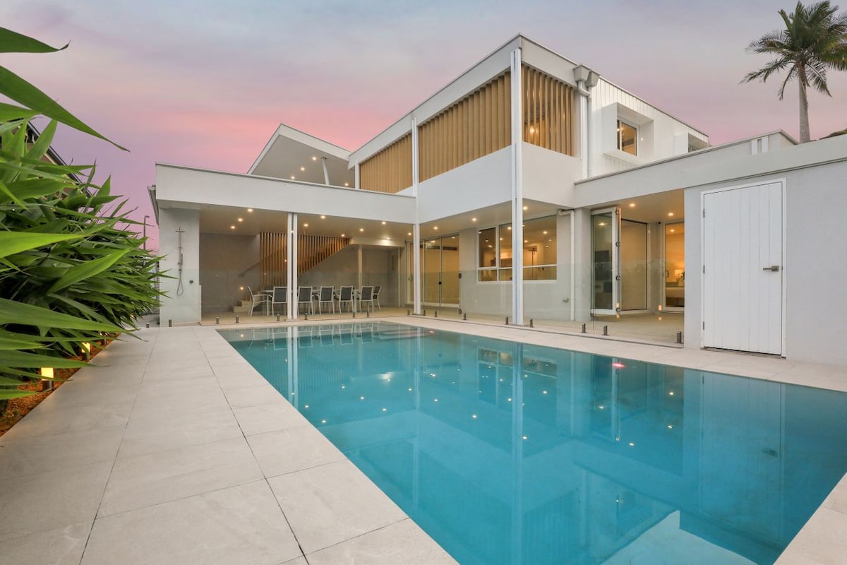 A modern villa is showcased with a clear swimming pool reflecting the colors of the sunset. Surrounding greenery enhances the serene environment. The expansive glass windows and doors of the house create a seamless connection between indoor and outdoor spaces.