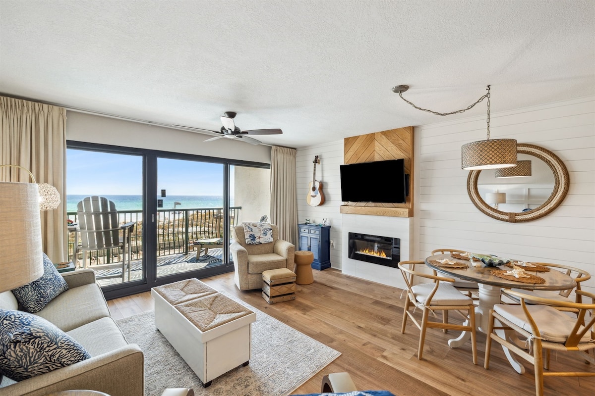 The living area features a modern coastal design with large glass doors that open to a balcony offering Gulf views. A comfortable sofa, accent chair, and a stylish coffee table are arranged around a cozy electric fireplace. A dining table with four chairs sits nearby, illuminated by natural light.