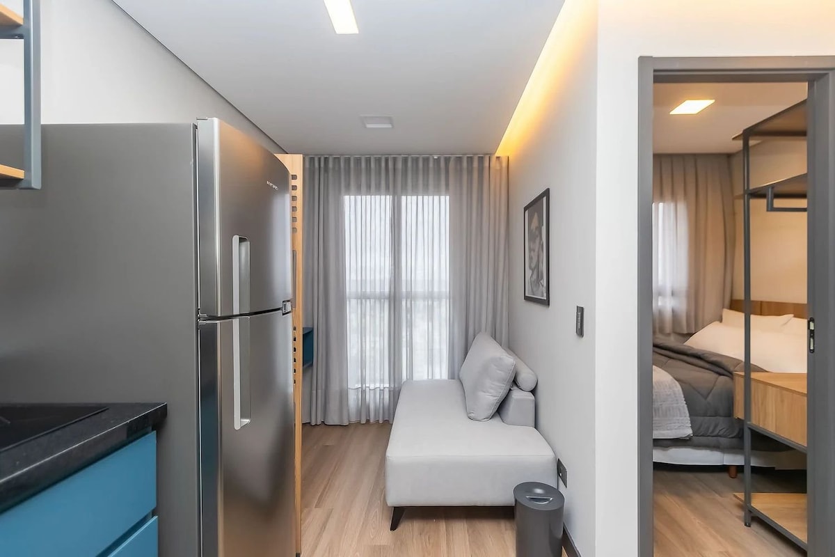 The image depicts a modern studio apartment with an open layout. A compact kitchenette with sleek cabinetry is visible to the left, while a light grey sofa is positioned against the wall. Large windows with sheer curtains allow natural light to illuminate the space, leading to a separate sleeping area.