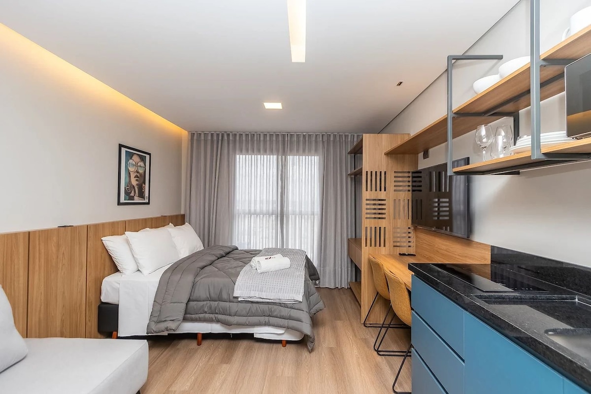 A modern studio space features a spacious bed with neutral-colored bedding, positioned against a wood-paneled wall. Natural light filters through sheer curtains, complementing the light-colored flooring. A compact kitchen area with sleek cabinetry and dining table is visible, creating an efficient layout.