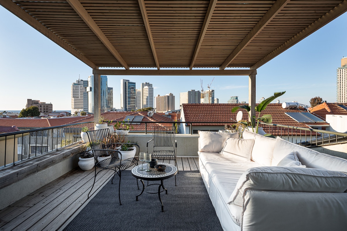An expansive rooftop terrace is featured, showcasing a comfortable outdoor lounge area with a large white sofa and a round table. Potted plants add greenery, while a view of the city skyline is visible in the background under a clear blue sky.