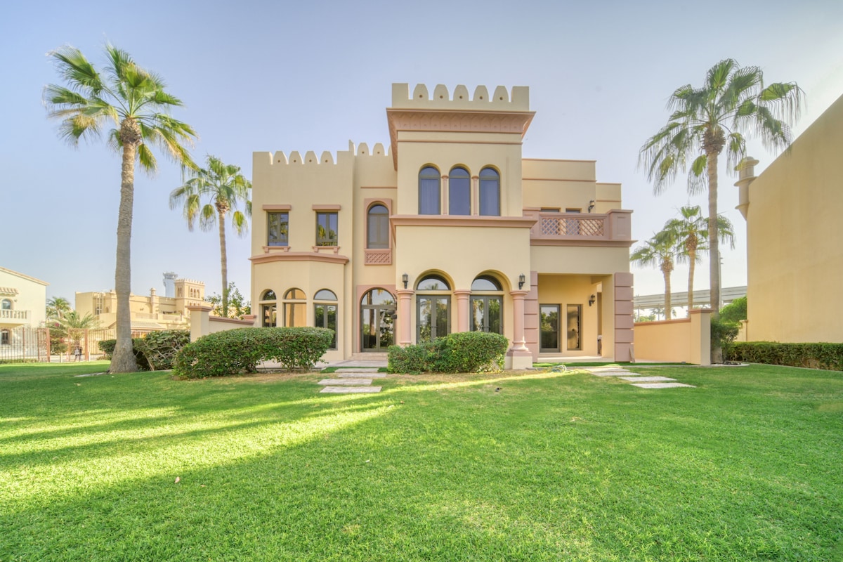 The exterior of the villa displays a distinctive architectural style, featuring tall arched windows and a prominent rooftop. Lush green grass surrounds the property, and palm trees offer a tropical ambiance. Pathways lead to the main entrance, inviting guests into the spacious outdoor area.