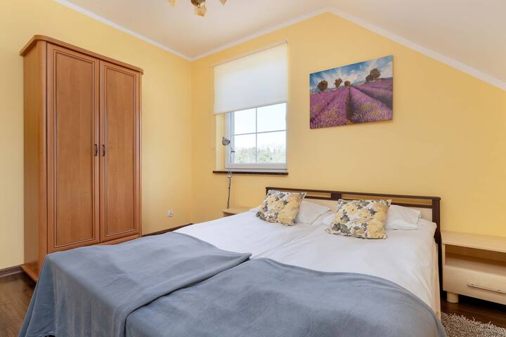 Спалня с голямо легло, светло спално бельо и шарено покривало. Стаята има и дървен гардероб и прозорец с изглед навън.