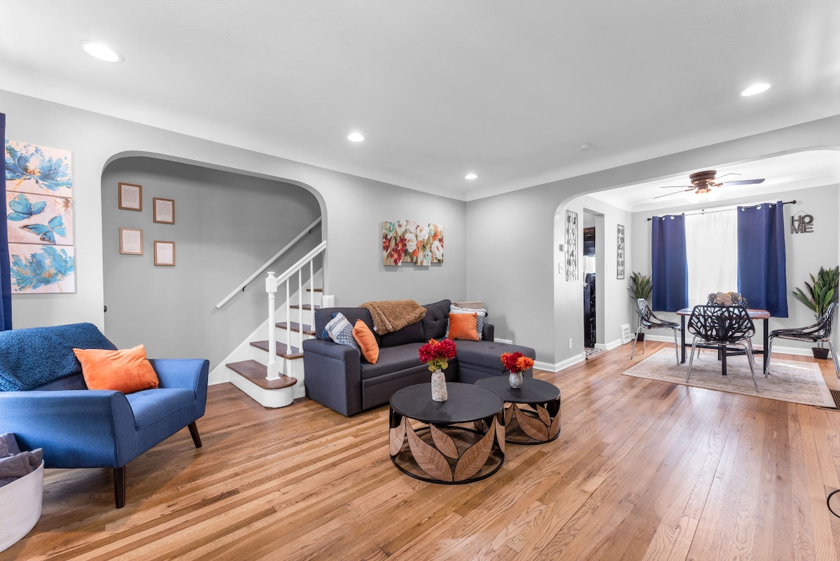 The living space features an open layout with polished hardwood floors and comfortable seating. An L-shaped sofa is positioned near a stylish coffee table, while a colorful wall art piece adds visual interest. A dining area is visible through an archway, with natural light streaming in from large windows.
