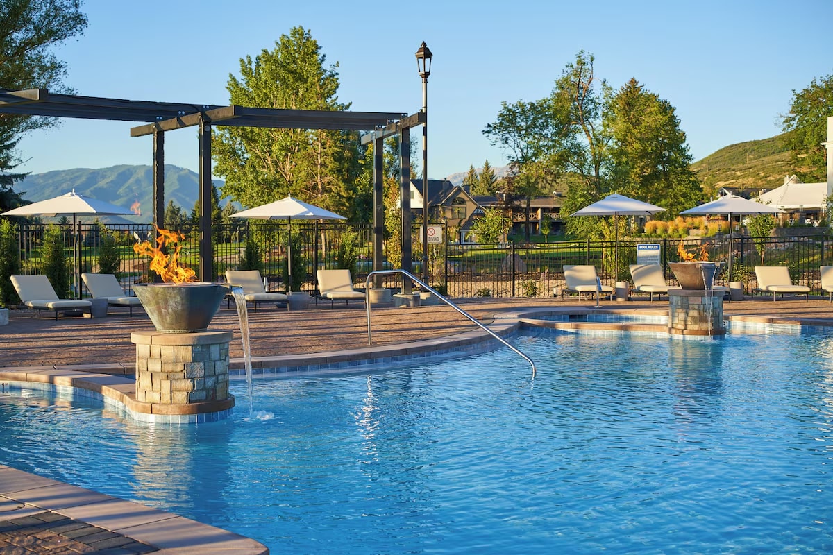An outdoor pool area features a clear, inviting pool with steps leading in. Surrounding the pool are lounge chairs and shaded umbrellas, providing comfort. Lush greenery and distant mountains are visible in the background, enhancing the serene ambiance of the space.