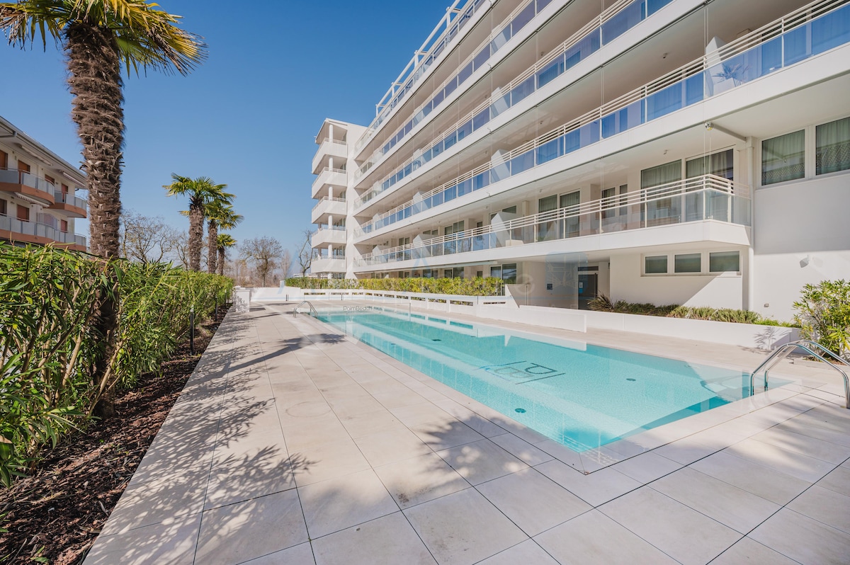 A modern building features a sleek swimming pool surrounded by a tiled deck, with landscaped greenery lining the perimeter. Tall palm trees are visible, adding a touch of nature to the contemporary setting. The clear blue sky enhances the inviting atmosphere of the space.