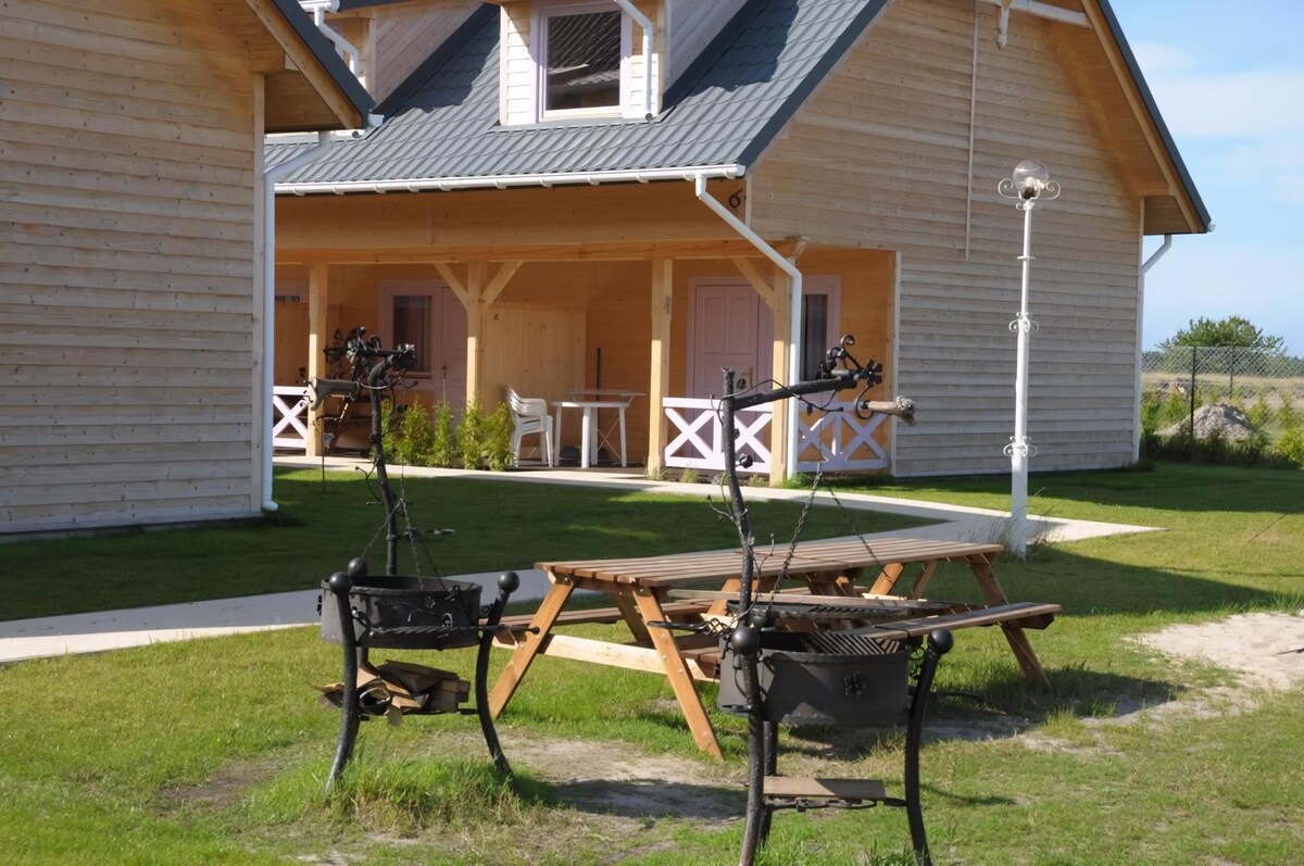 A well-maintained outdoor area is presented, featuring two barbecue grills positioned on a grassy lawn. A picnic table is situated nearby, providing a space for outdoor dining. The wooden two-story building is visible in the background, highlighting a welcoming and natural environment.