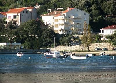 A view of the waterfront shows several boats docked in a calm bay. In the background, buildings line the hillside, surrounded by greenery. The scene reflects a tranquil day by the water, with the soft ripples of the sea visible in the foreground.