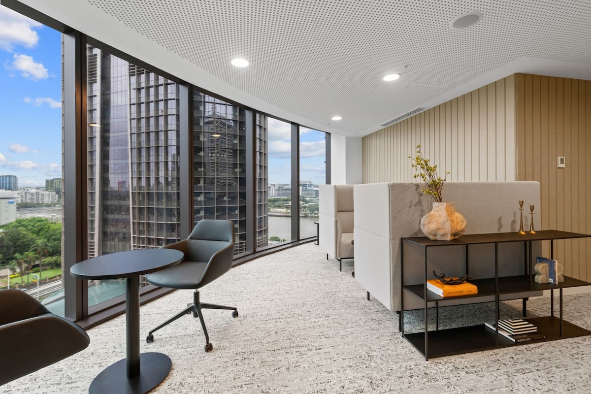 A bright lounge area features large windows showcasing city views. Modern furnishings include a round table with a black surface, a comfortable chair, and two plush seating units. The space is adorned with decorative elements like vases and a sculptural piece, creating a relaxed atmosphere.
