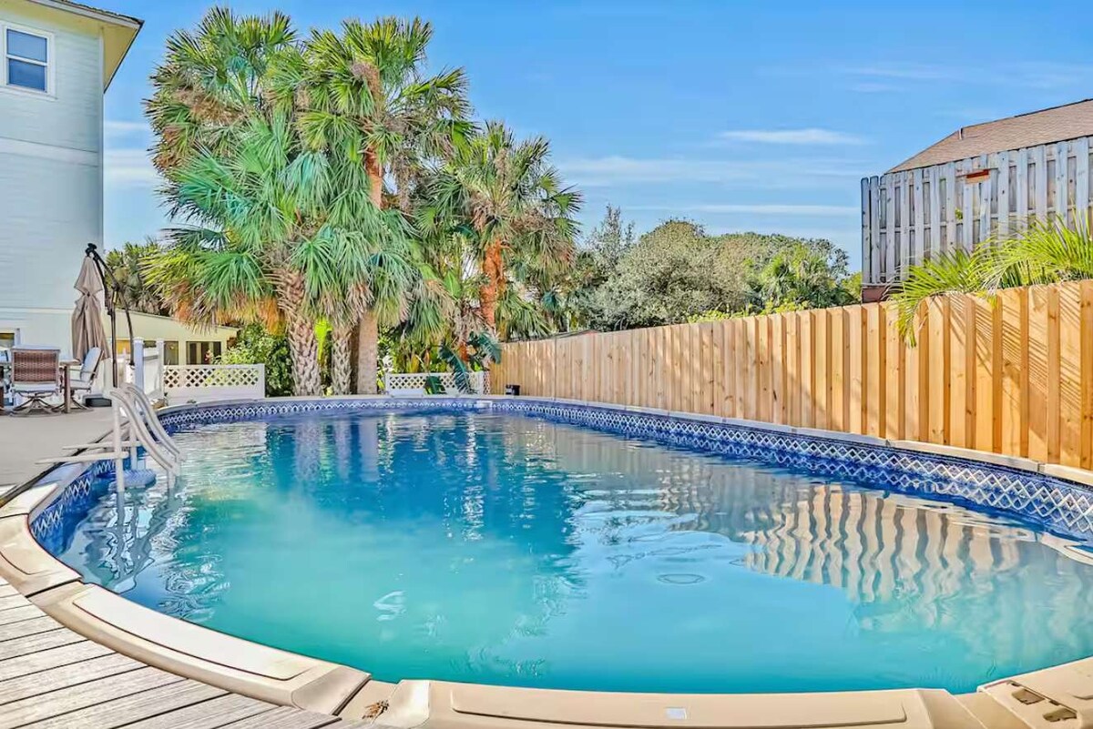 A private in-ground pool is surrounded by tropical palm trees and a wooden fence. Poolside lounge chairs are positioned for relaxation under a clear blue sky, with the shimmering water reflecting the sunlight.