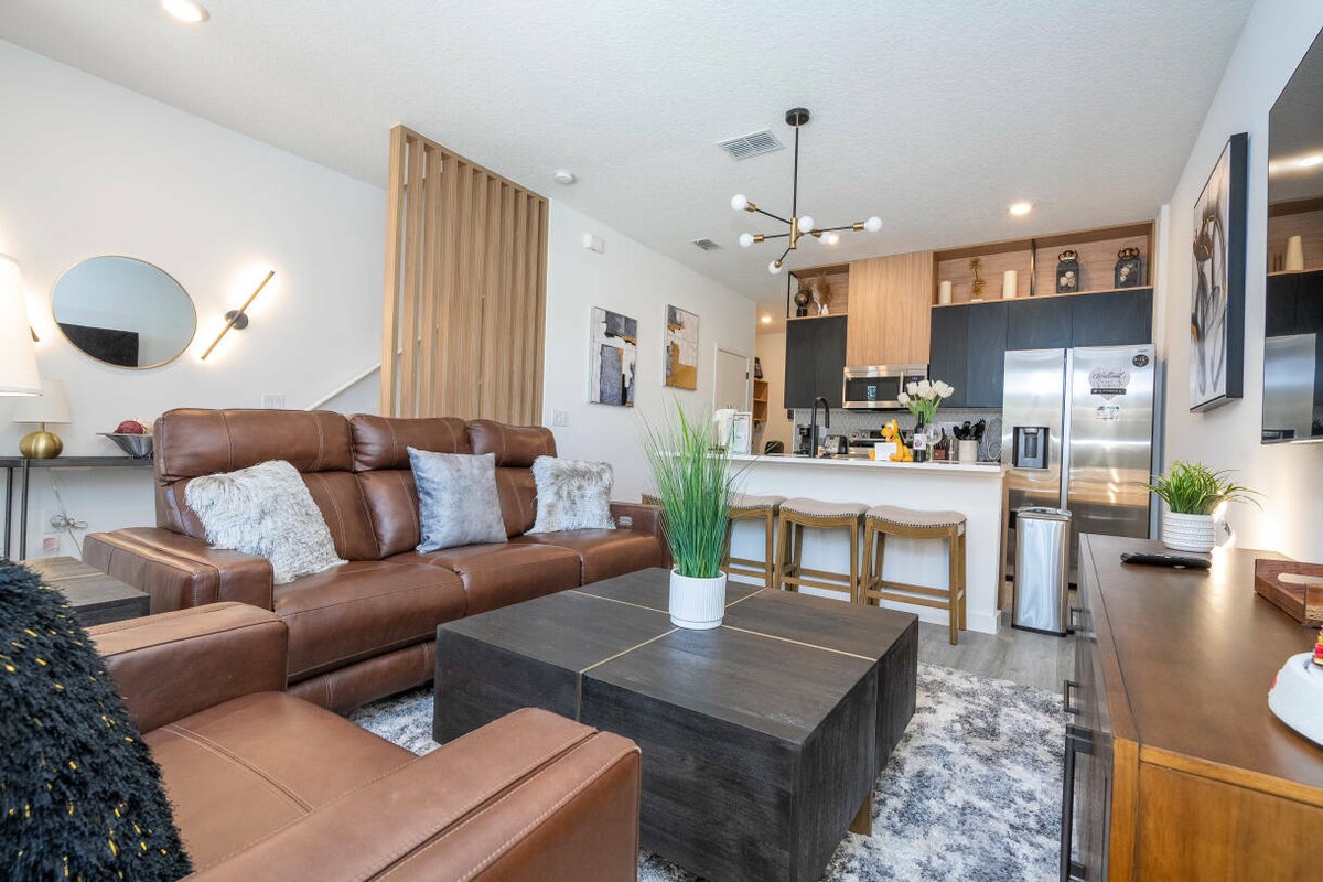 A cozy living area showcases a leather sectional sofa adorned with decorative pillows. A wooden coffee table sits at the center, complemented by a lush green plant. The open kitchen features modern appliances, bar stools, and a stylish light fixture above, creating a functional yet inviting space.