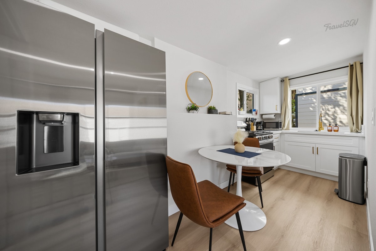 The kitchen area showcases modern appliances including a stainless steel refrigerator and a gas stove. A round dining table with one chair is positioned near a window, allowing natural light to illuminate the space. Minimalist decor elements add a touch of warmth.