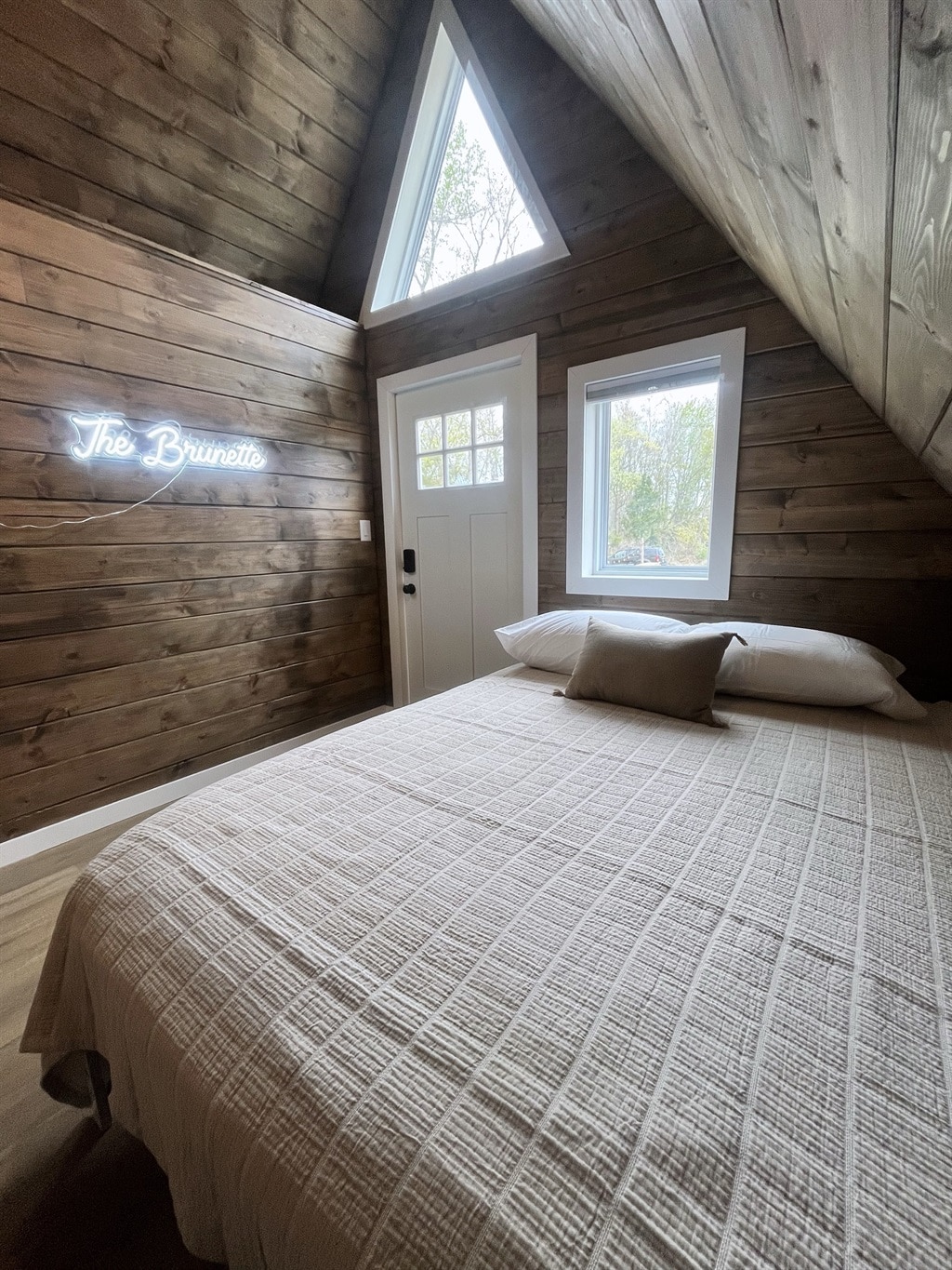 A cozy sleeping area features a spacious bed dressed in a textured, neutral quilt. Natural light filters through two windows, illuminating the wooden walls and triangular ceiling. A subtle sign reading 'The Brunette' adds a touch of charm to the inviting space.