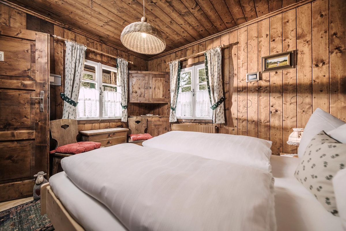 The bedroom features wooden paneling and a warm, natural color palette. A spacious double bed is dressed in white linens, and two windows with lace curtains provide natural light. A rustic light fixture hangs above the bed, while a wooden dresser and a small desk complete the cozy atmosphere.