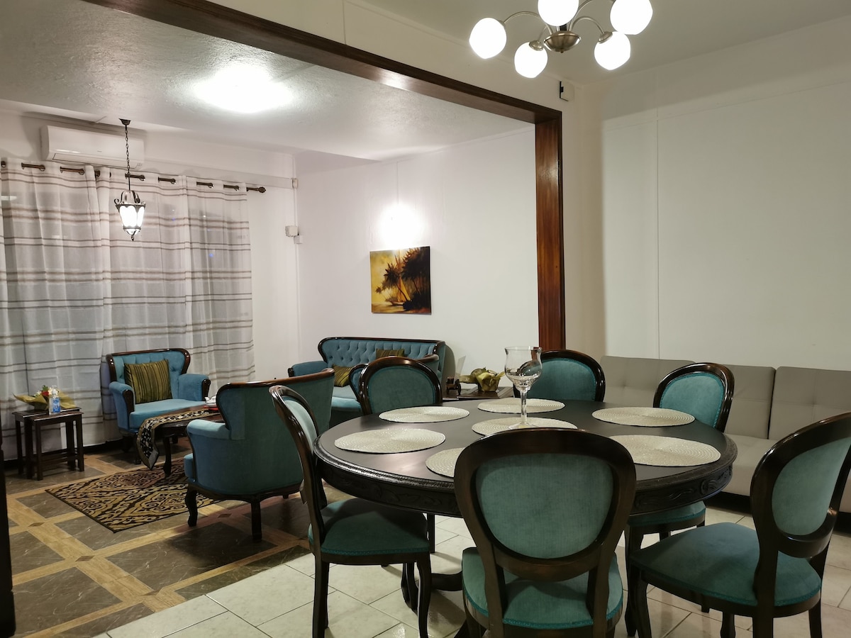 An open living and dining area showcases a round dining table surrounded by teal upholstered chairs. A seating arrangement consisting of a light grey couch and teal chairs is visible. Natural light enters through large windows covered with white curtains, creating a welcoming ambiance.