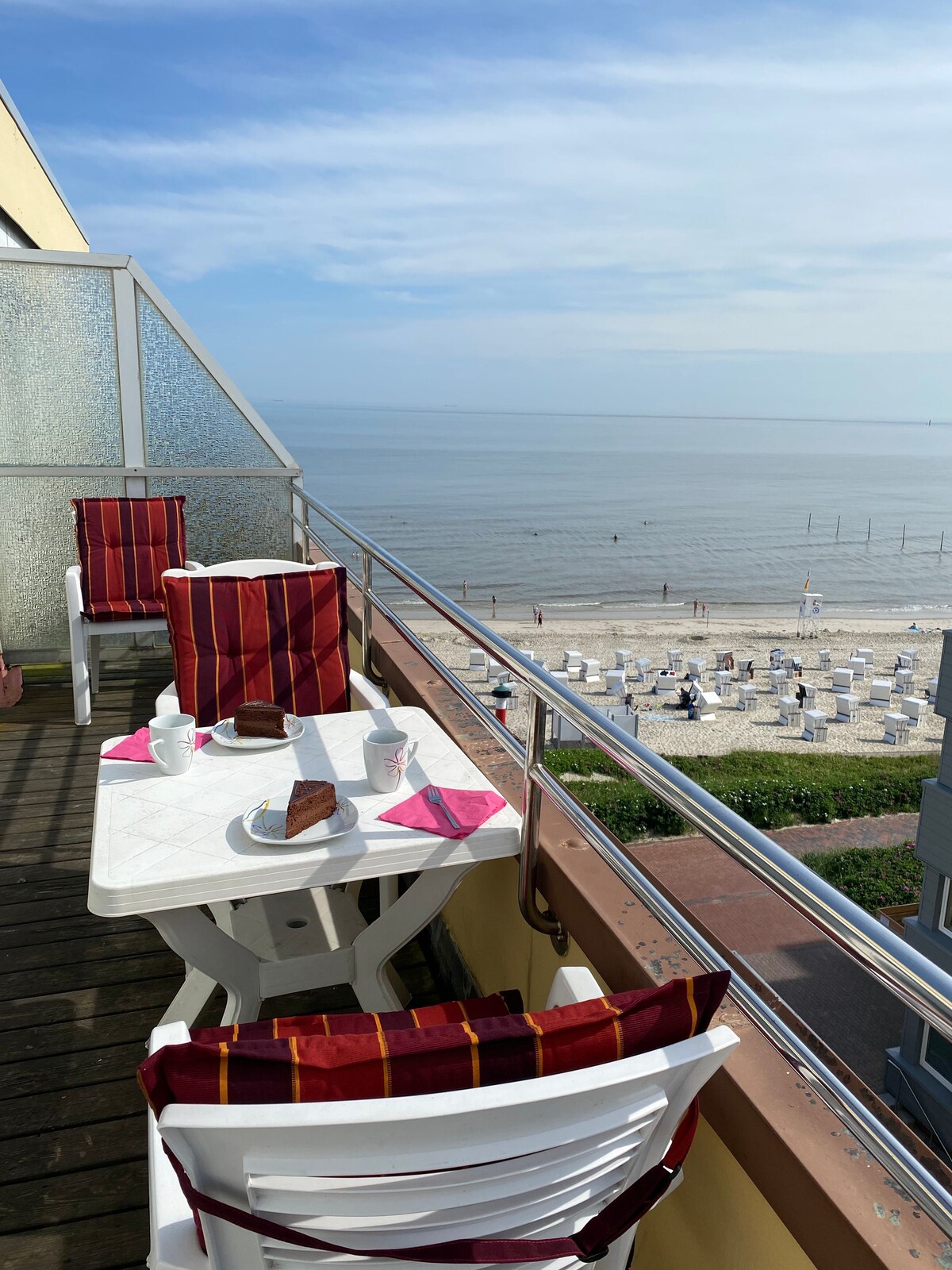 A rooftop terrace is featured with a small table set for breakfast, including two cups and a plate with cake. Two chairs with red-patterned cushions are positioned beside the table, offering a direct view of the beach and sea, creating a serene outdoor dining space.