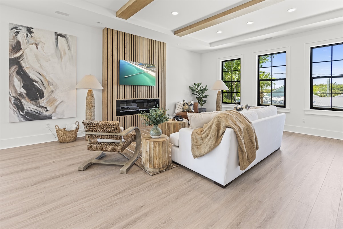 A spacious living area is featured, with a large white sofa and wooden accent chairs. A decorative lamp stands beside the sofa. Wooden beams are visible on the ceiling, while a large artwork and a media unit accent the room. Natural light enters through multiple windows.
