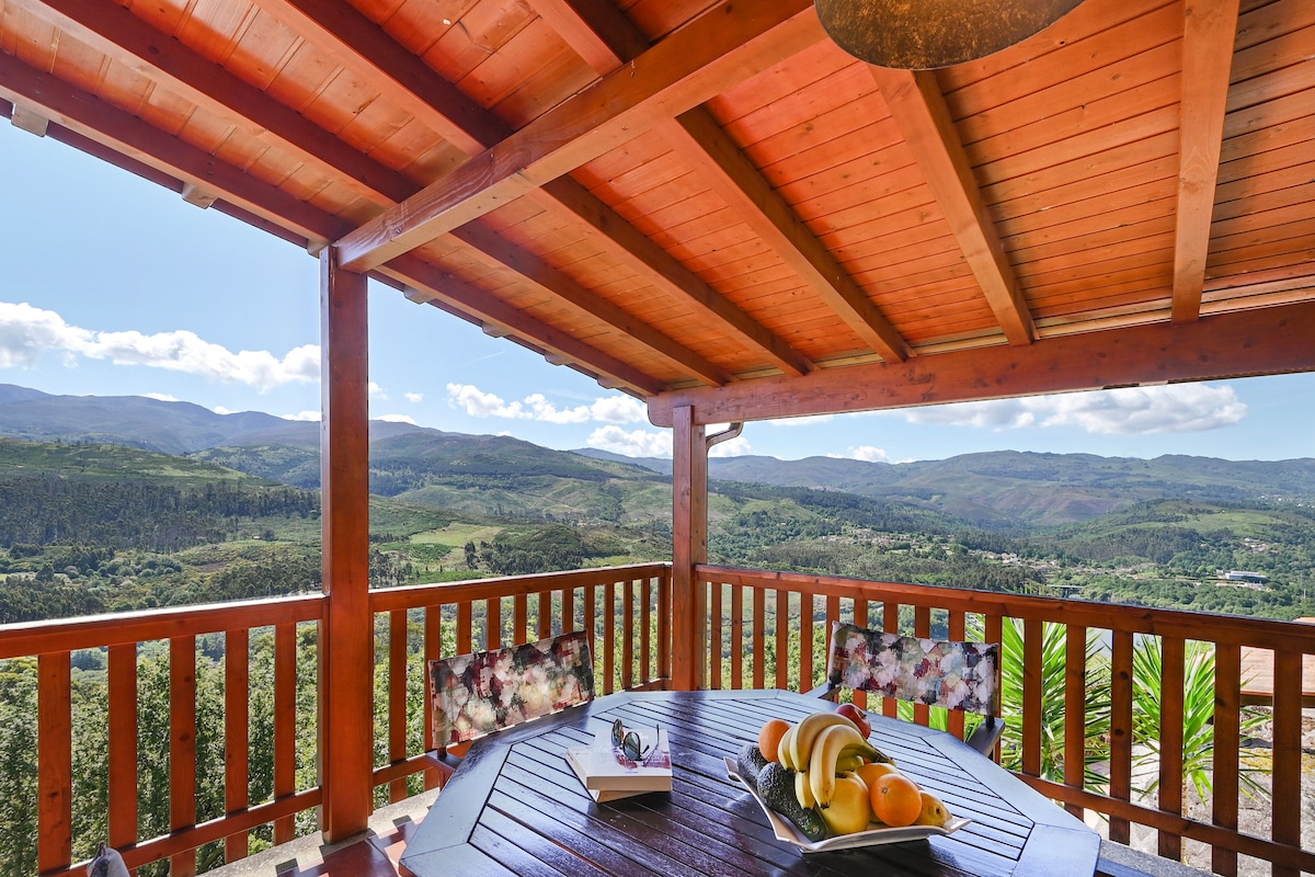A covered outdoor terrace is featured, furnished with a round wooden table displaying an arrangement of fresh fruits. Plush cushions adorn the wooden chairs, while a scenic view of rolling hills and valleys stretches in the background under a clear blue sky.