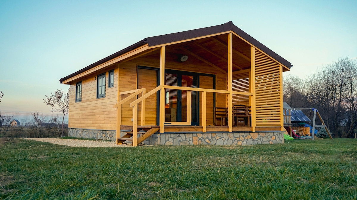 A wooden cottage features a spacious covered porch, furnished with seating for outdoor relaxation. Large windows allow for natural light, and the blend of wood and stone enhances the charming exterior. A grassy area surrounds the cottage, providing a serene environment.