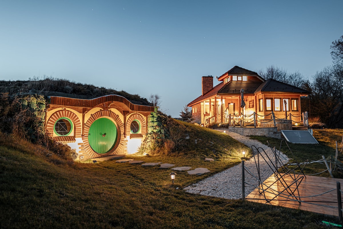 A Hobbit-style house is depicted with a unique round green door, complemented by warm outdoor lighting. The pathway leads from the house to the charming entrance. The structure showcases natural wooden elements and is set against a twilight sky, surrounded by gentle terrain.