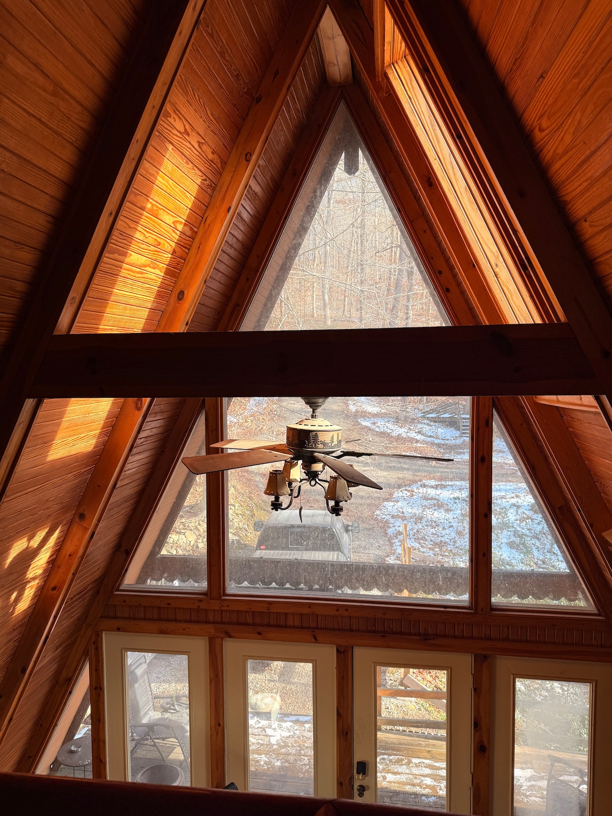 An A-frame cabin interior is highlighted, showcasing high wooden ceilings with large windows that allow natural light to fill the space. A ceiling fan is prominently displayed, and a glimpse of the outdoors reveals a serene wooded area with patches of snow.