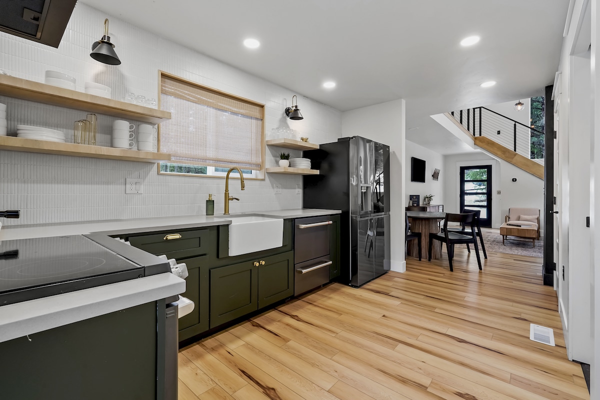 A modern kitchen features dark cabinetry and open shelving displaying dishes. A farmhouse sink is placed below a window, while stainless steel appliances stand in contrast. Light wood floors run throughout, connecting to a dining area visible in the background with a glimpse of the living space.
