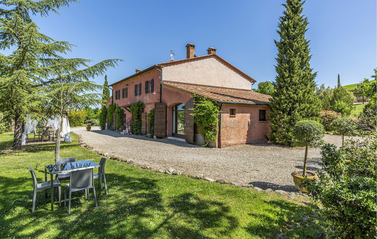 A charming country house, characterized by a warm terracotta exterior, is surrounded by lush greenery and Mediterranean plants. The outdoor area features a gravel pathway leading to the entrance, as well as a shaded dining table accompanied by modern chairs, ideal for gatherings.