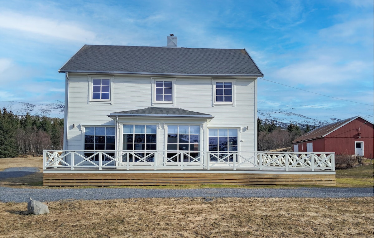 A two-story house is displayed, featuring a light-colored exterior and multiple large windows. A spacious wooden terrace encircles the house, while a gravel pathway leads to the entrance. The backdrop includes gentle slopes and distant mountains under a clear blue sky.