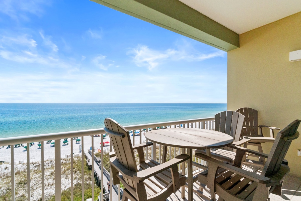 A private balcony overlooks the Gulf, featuring a round wooden table accompanied by four adirondack chairs. The calm blue waters and sandy beach are visible in the distance, creating a serene coastal setting.