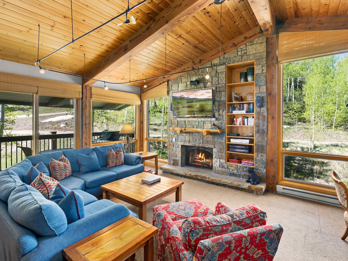 A spacious living room showcases a vaulted ceiling with pine beams, enhancing the airy feel. A mountain stone gas fireplace serves as a focal point, flanked by built-in shelves. Soft seating arrangements include a blue sectional and patterned armchairs, with large windows offering views of the outdoors.