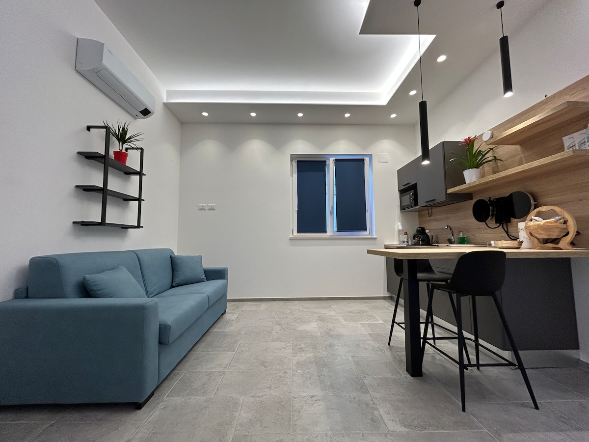 A light-filled living space features a blue sofa and a kitchenette with modern fixtures. The wooden countertop is complemented by a black bar stool, with small decorative plants enhancing the area. Bright lighting fixtures and an air conditioning unit create a comfortable atmosphere.