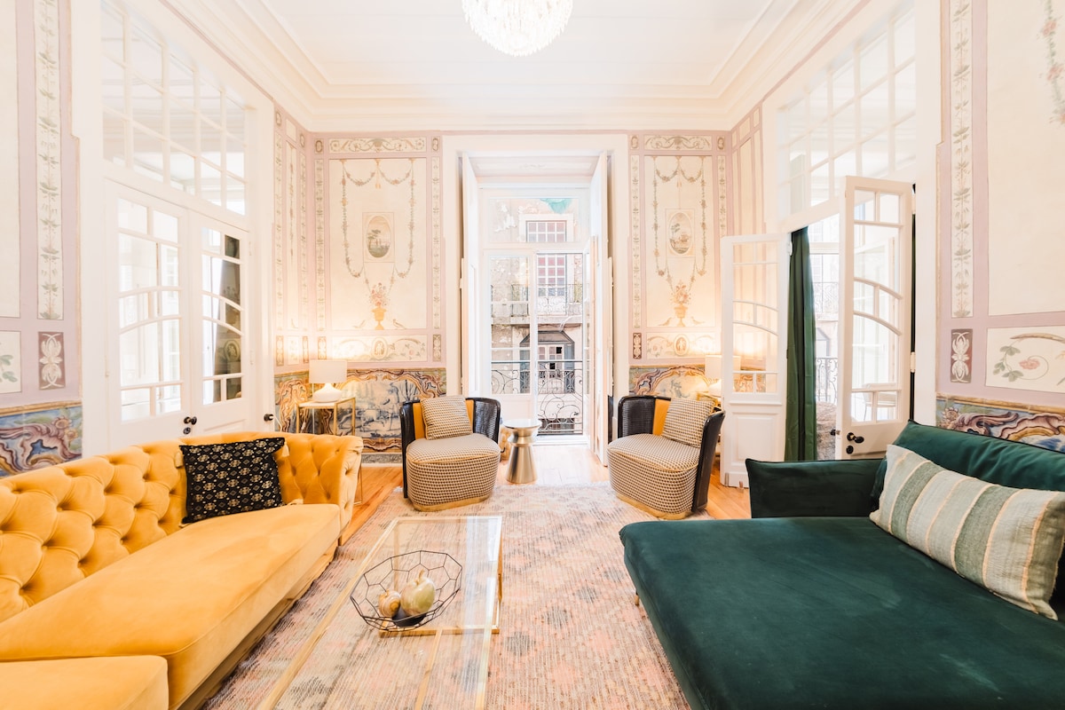 A spacious living area showcases elegant hand-painted walls, with two plush armchairs and a large green sofa offering comfortable seating. A light fixture hangs from the ceiling, and a glass coffee table is placed on a textured area rug. Natural light is allowed through large open doors.