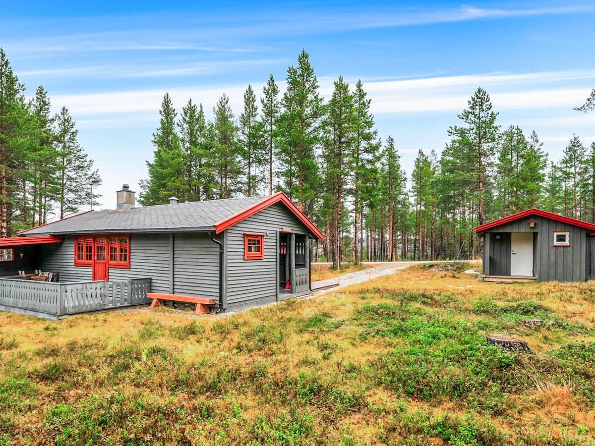 A cozy holiday home is set within a natural landscape, surrounded by tall green trees. The building features a grey exterior with red accents, including window frames and doorways. A spacious wooden deck is visible in front, along with a separate building nearby.