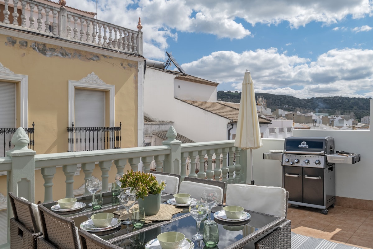 An outdoor terrace is presented with a dining table set for six, featuring plates and glasses. A grill is visible in the corner, accompanied by an umbrella providing shade. The backdrop includes rooftops and distant mountains, under a partly cloudy sky.