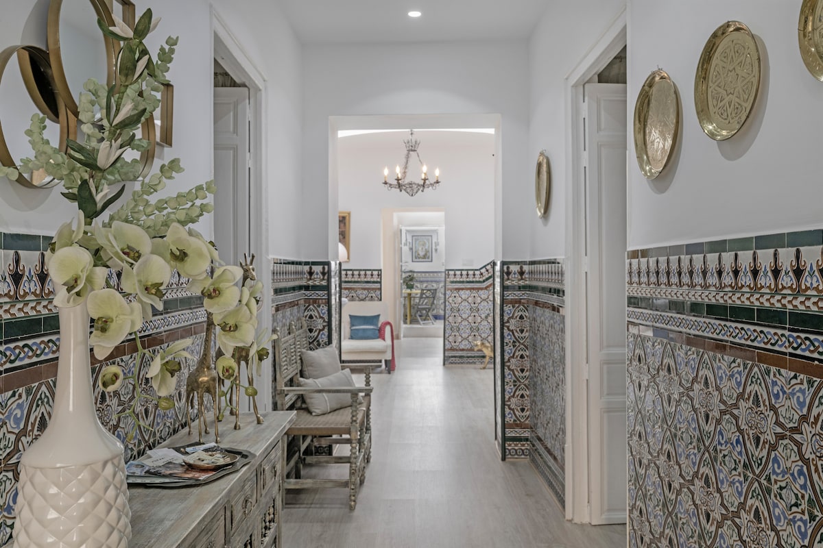 A bright corridor features intricate tile work along the walls, complimented by decorative plates. A console table adorned with a vase of flowers stands to one side, while artwork can be seen at the far end, leading to a welcoming, open living space.
