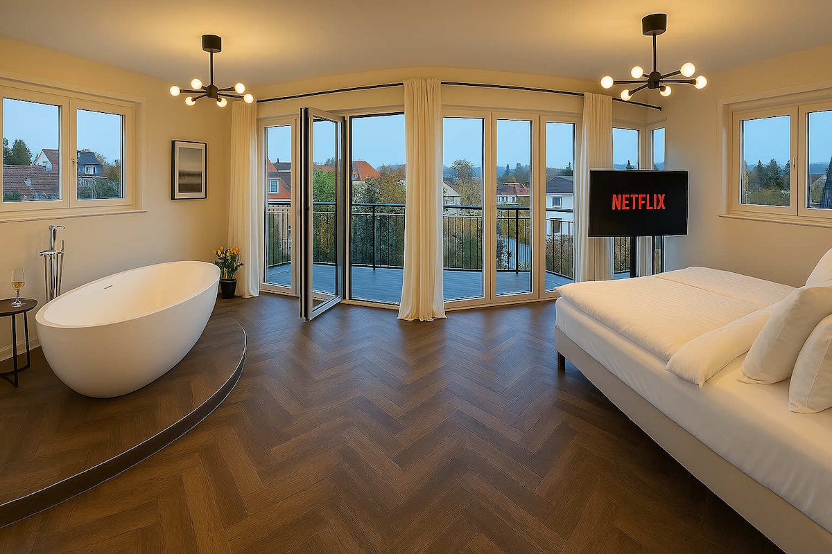 The spacious bedroom features a large window wall that opens to a balcony, allowing for ample natural light. A modern freestanding bathtub and a 75" TV are present, with neutral-toned furnishings arranged to create an inviting space.
