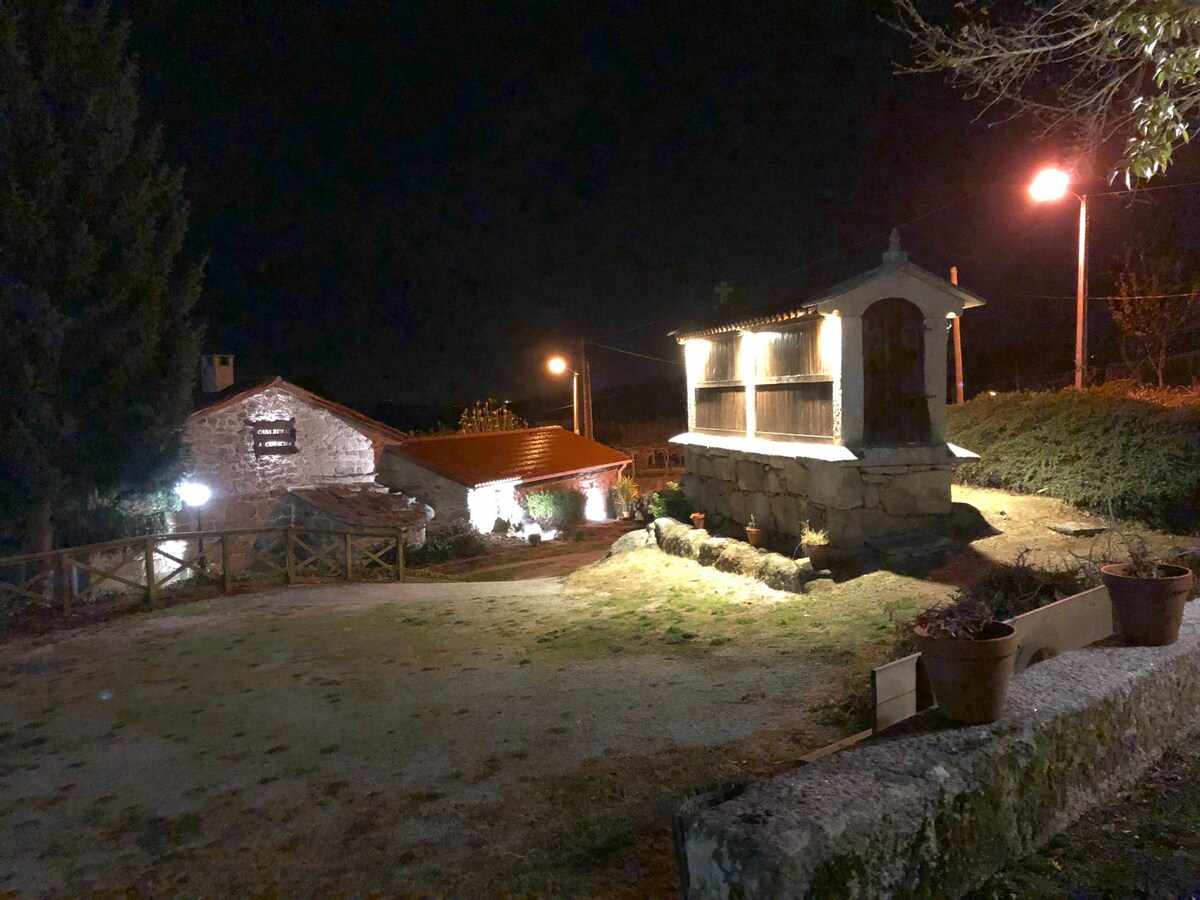 The property is illuminated softly at night, showcasing a rustic house alongside a traditional stone structure. A well-maintained garden area is visible, with pathway lights guiding the way. Potted plants sit on the stone wall, contributing to the serene atmosphere.