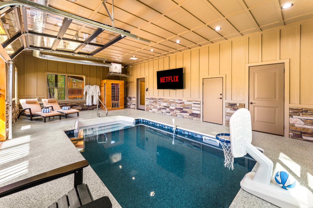 An indoor heated pool is surrounded by a spacious area, featuring lounge chairs and wall-mounted screen displaying Netflix. The pool area is enhanced by wooden accents and large doors that can be opened, providing light and a connection to the outdoors.