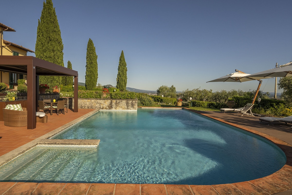 A spacious swimming pool with a stone edge is surrounded by manicured gardens. The pool features a Roman staircase for easy access and is bordered by parasols and sun loungers. Lush greenery and olive trees create a serene backdrop to the outdoor space.